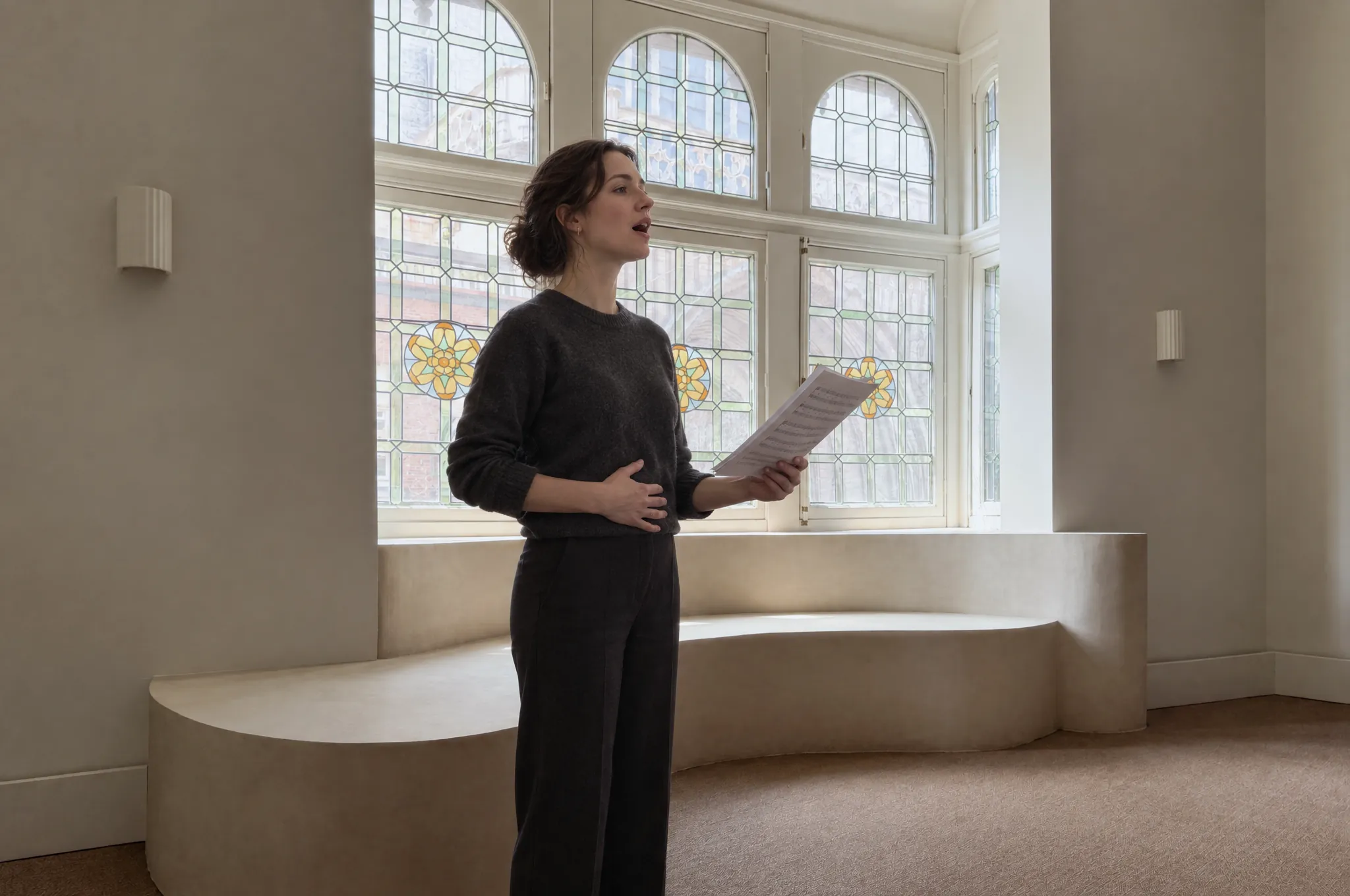 Een volwassen vrouw zingt met de hand op haar buik voor ademsteun, staand bij de glas-in-loodramen van de salon